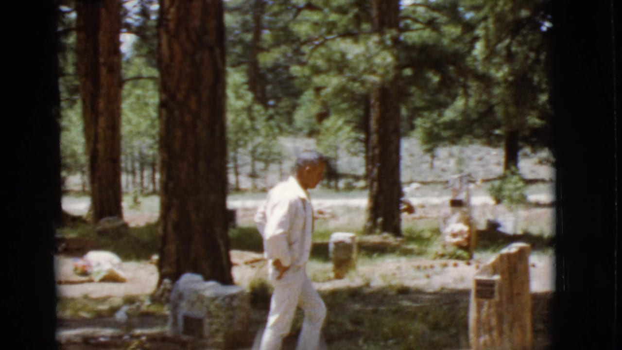 Man walking beside a headstone in a wooded cemetery clearing.
