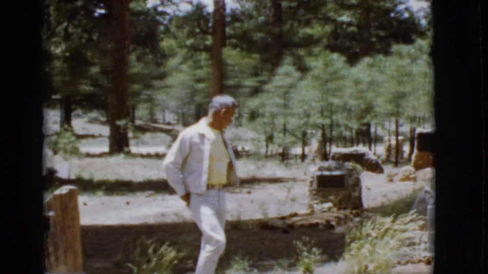 At the end of the clip, the man passes a foreground marker while sunlit trees and cemetery stones remain visible behind him.
