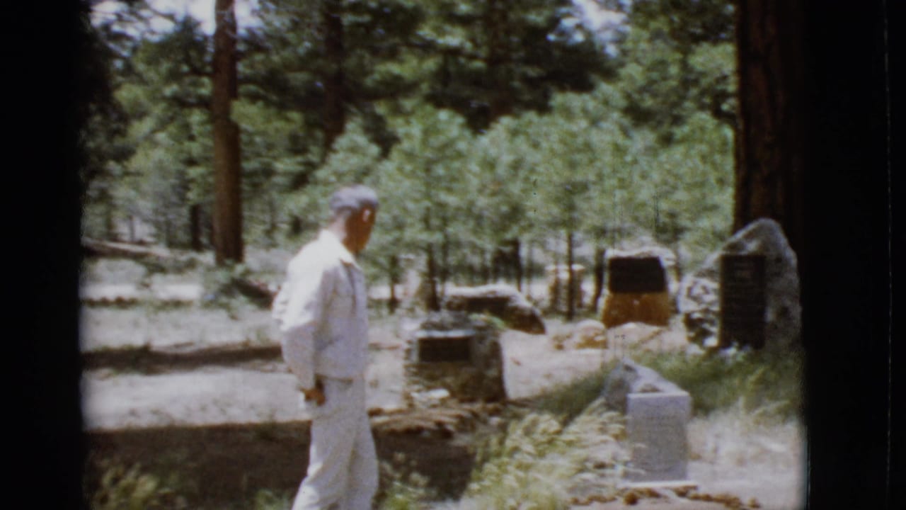 Man in a white suit walks through a pine forest toward stone markers.