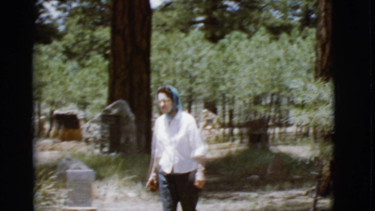 Woman in a white blouse and blue headscarf walking near gravestones among pine trees.