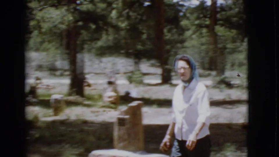 A soft 8mm frame shows the woman nearer the camera, with scattered gravestones and pine trees in the background.