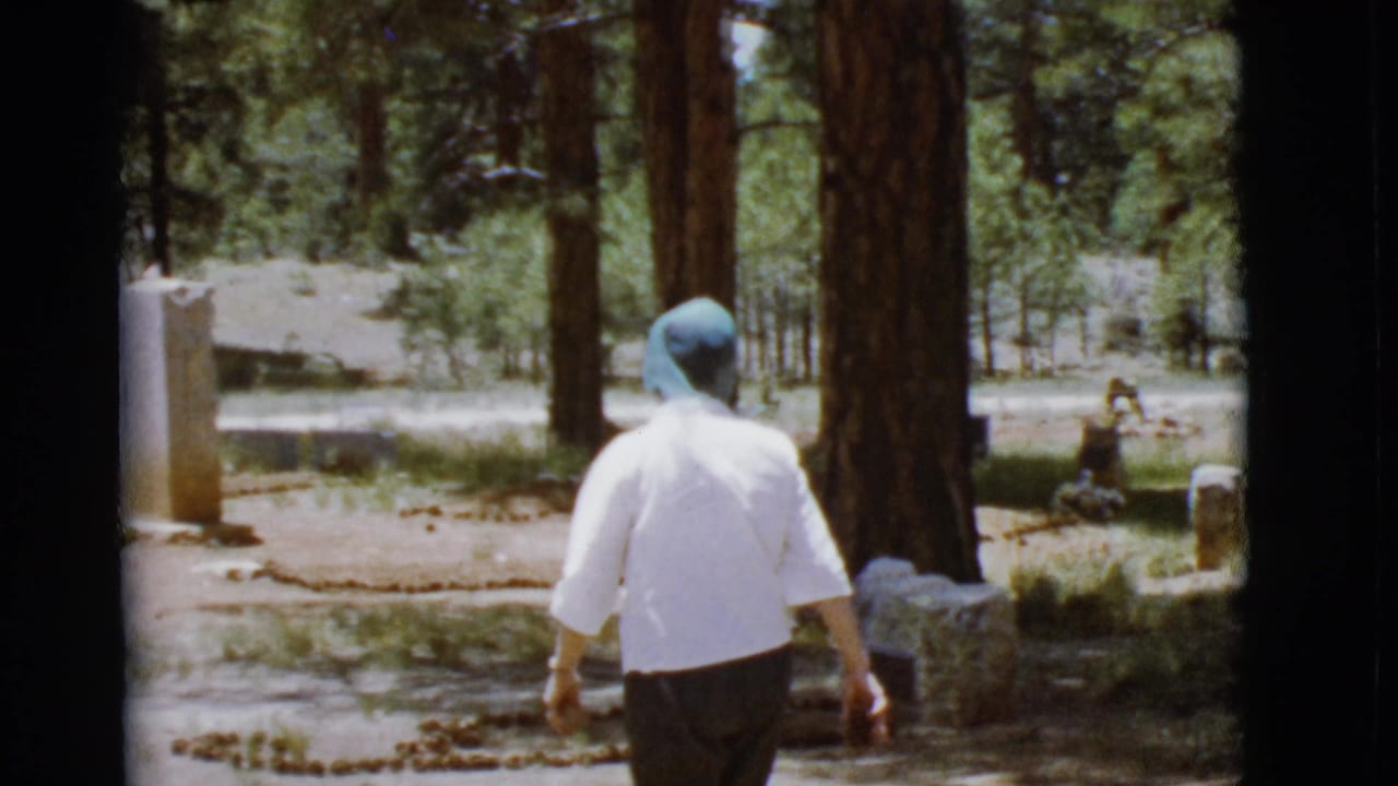 Woman in a blue headscarf walks through a wooded cemetery toward a grave marker.