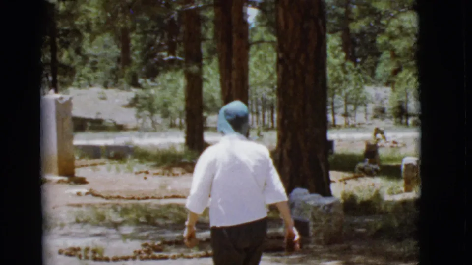 In 1968 Arizona 8mm footage, a woman in a white shirt and blue headscarf walks among trees toward a decorated grave area.