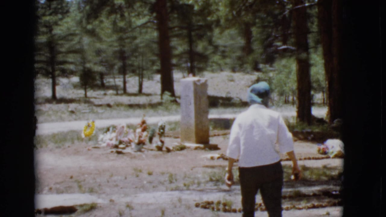 Woman stands near a decorated grave and stone marker in a wooded cemetery.