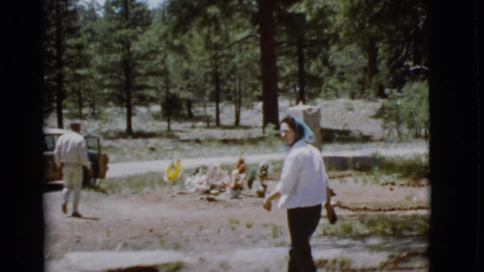 A man walks toward a brown parked car while the woman in a blue headscarf passes near a decorated grave in the wooded cemetery.