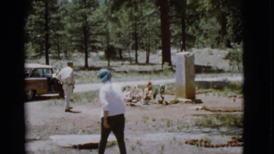 The woman crosses the foreground near the flowered grave while a man stands by a brown car in this 1968 Arizona cemetery scene.