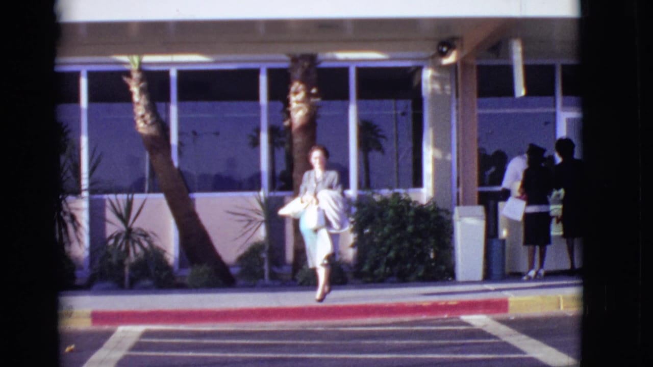 Woman in a grey dress walks near a red curb outside a glass-fronted building.