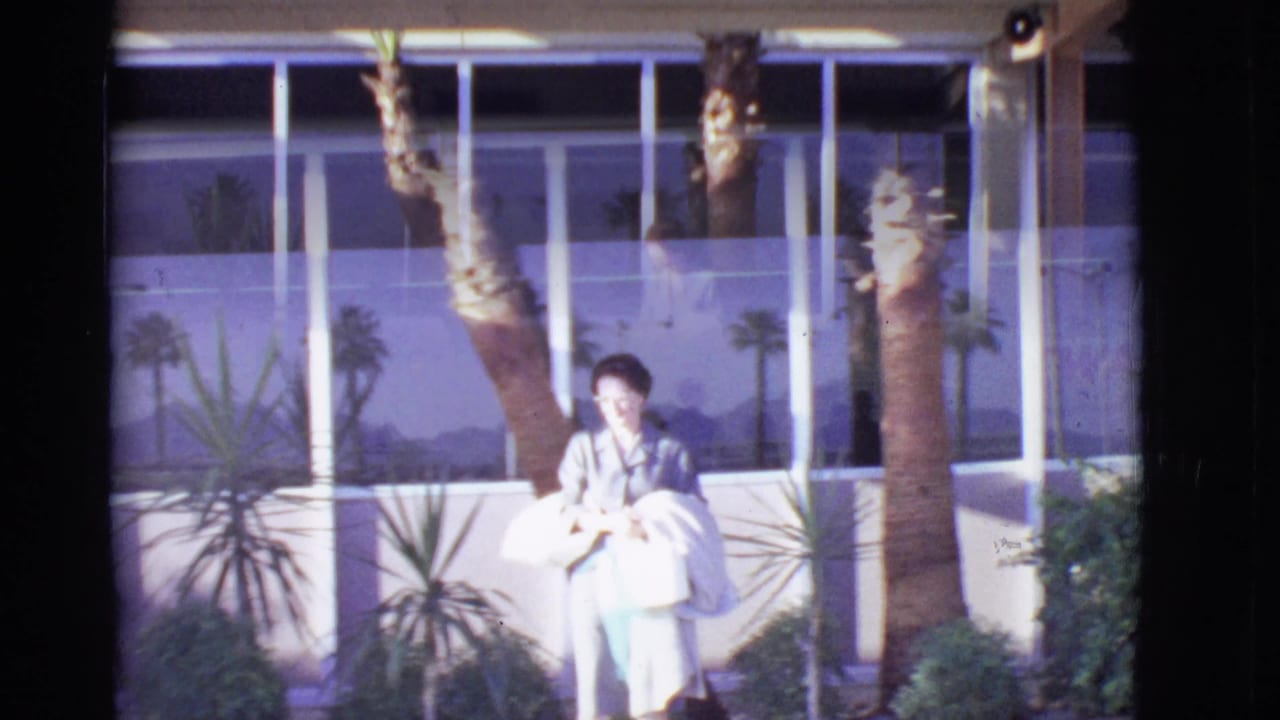 Woman stands or walks near palm trees and large windows outside a building.