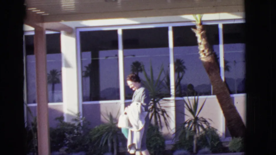 In Arizona 1968, the woman walks in profile beside palms, shrubs, and large reflective windows on a sunny exterior walkway.