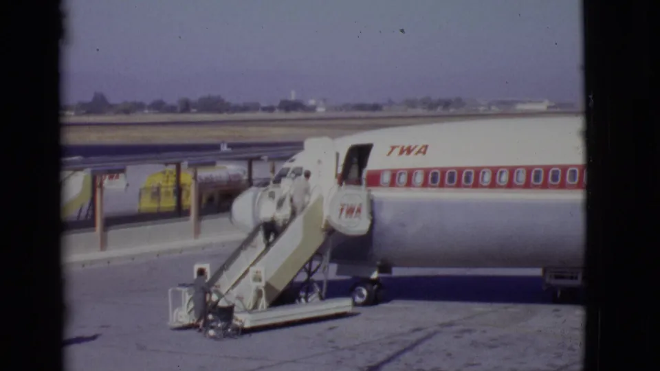 A TWA aircraft is parked beside mobile stairs as passengers continue boarding from the open tarmac area.