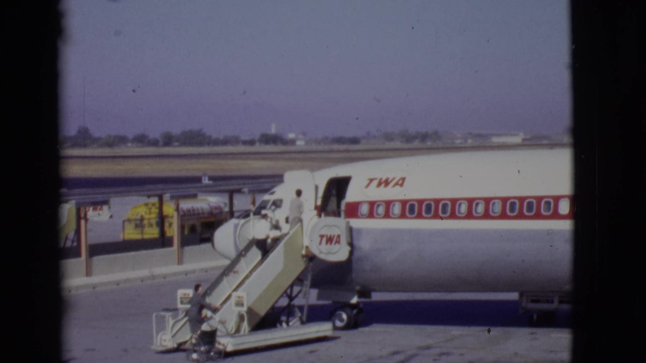 TWA airliner at an airport gate with mobile stairs and a yellow service truck nearby.