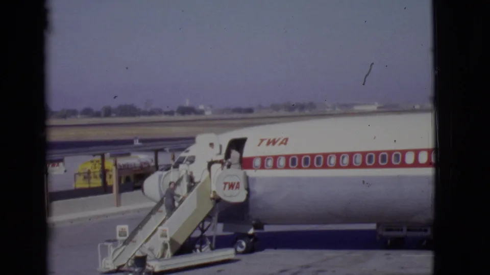 Late-1960s airport boarding view with a TWA airliner, mobile stairs, and a passenger nearing the open forward door.