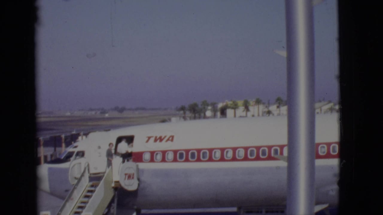 Passengers board a white TWA aircraft by mobile stairs on an airport tarmac.