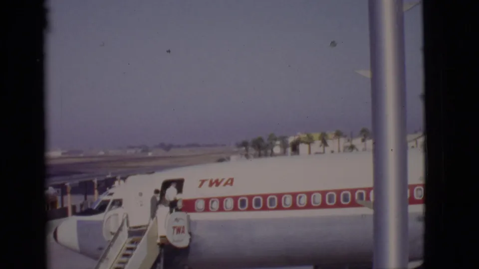Arizona, 1968 home-movie frame showing a TWA aircraft, red window band, mobile stairs, and passengers boarding under a clear sky.