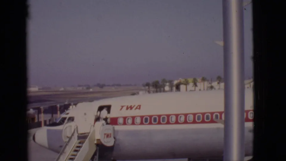 Arizona, 1968 frame of a TWA aircraft with passengers near the mobile stairway, palm trees beyond the tarmac, and a vertical obstruction at right.