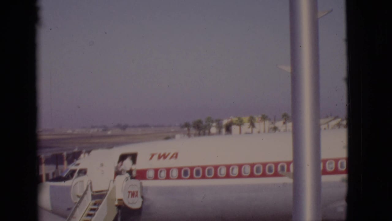 TWA jet at an airport gate with mobile stairs and a person near the fuselage.