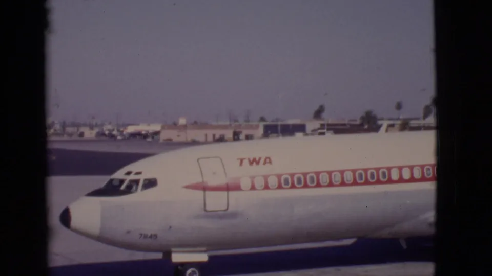 Home-movie close-up of a TWA jet's forward fuselage at an Arizona airport in 1968, with ramp buildings in the background.