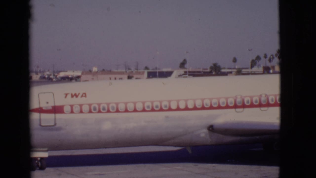 TWA aircraft fuselage seen broadside with airport buildings behind it.