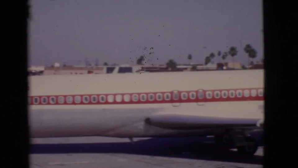 Home-movie frame showing the midsection of a TWA aircraft, red window stripe, and engine area as it taxis past.