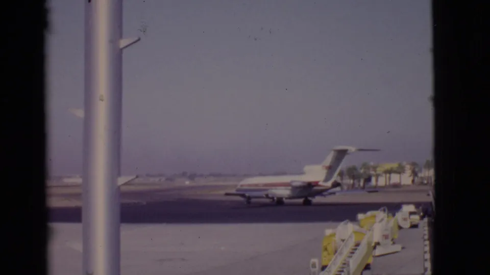 A soft 8mm frame shows the TWA jet lower in the scene, partly framed by a window post and open airport tarmac.