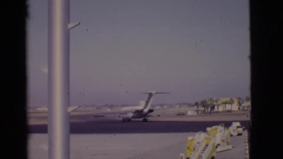 A large passenger airplane taxis across an Arizona runway in 1968, viewed through glass with palm trees and yellow service equipment visible.