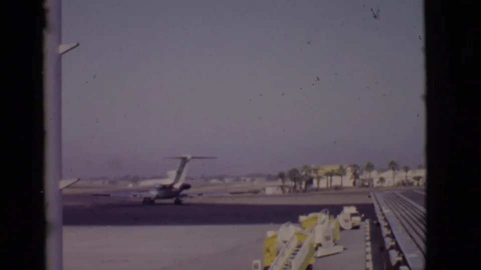 The 1968 Arizona home-movie frame shows a rear-engine passenger jet taxiing past ground equipment and palm trees from inside the terminal area.