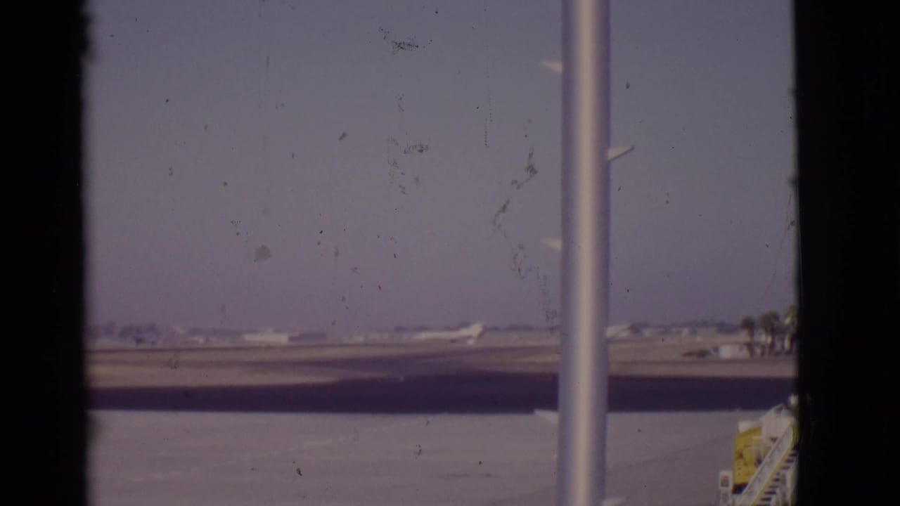 Distant white airplane on a runway seen through dark terminal window edges and a vertical pole.