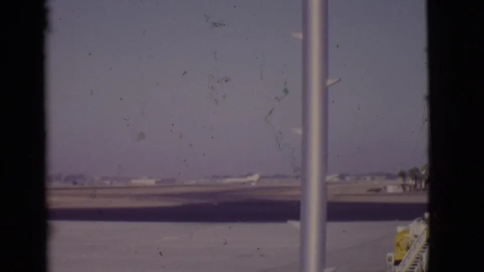 1968 Arizona 8mm frame showing a distant white airplane on a runway through terminal glass and dark window framing.