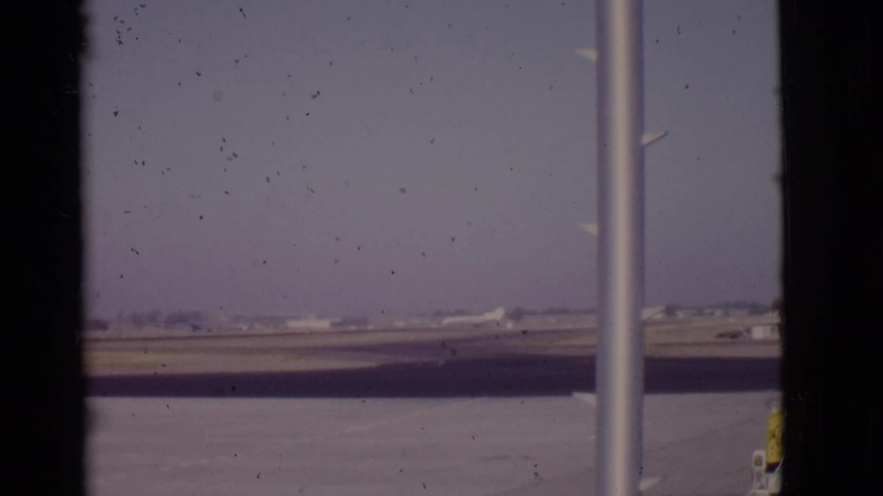 White airplane in the distance near the runway, framed by vertical poles and window edges.