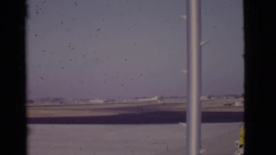 1968 Arizona airport view with a distant white commercial airplane beyond terminal window poles and a hazy runway.