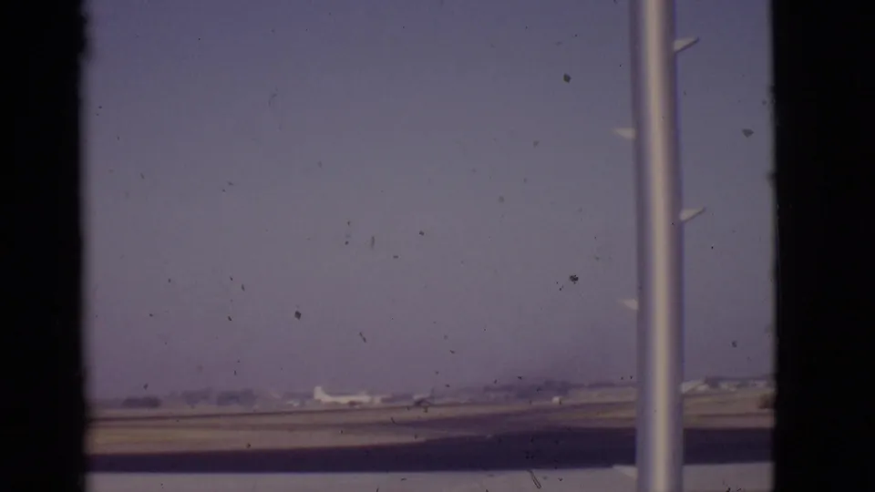1968 Arizona 8mm airport frame showing a runway, small distant airplane, hazy sky, and terminal window pole.
