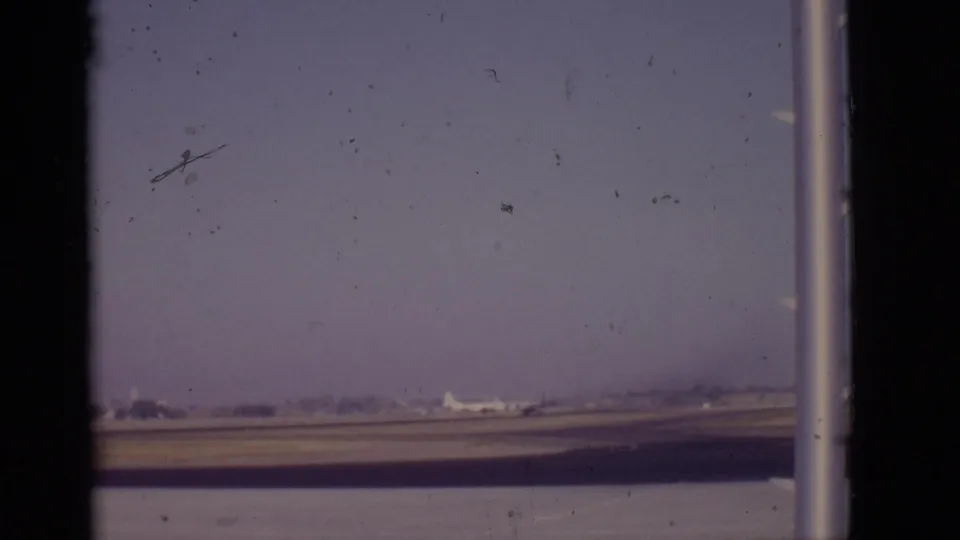 1968 Arizona runway view with a distant white aircraft, terminal window supports, muted color, and visible 8mm film wear.