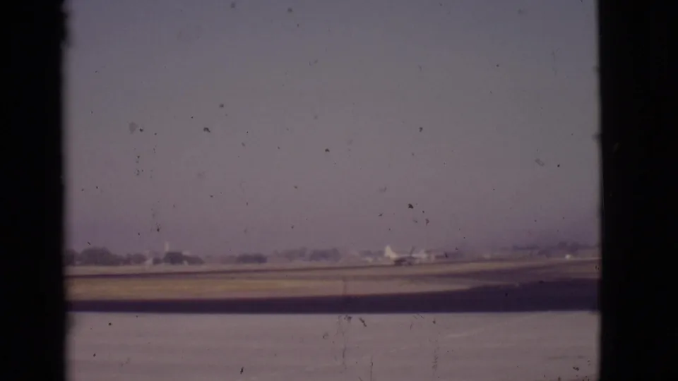 Distant commercial airplane on an Arizona runway in 1968, partially divided by a dark vertical window frame and soft film grain.