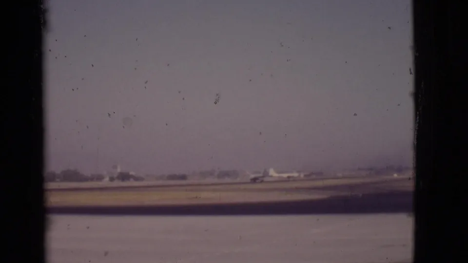 1968 8mm view of a small distant airplane on an Arizona runway with muted color, dust marks, and low airport buildings.