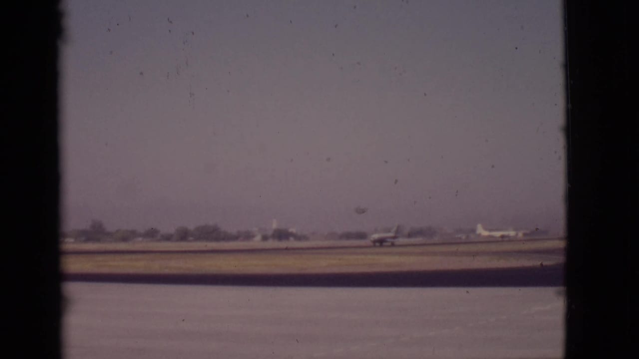 Propeller airplane on a runway beyond water, seen through dark frame edges under a hazy sky.