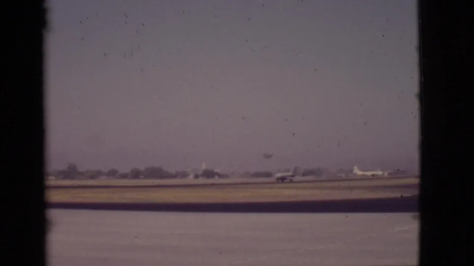 1968 Arizona 8mm frame showing a small propeller airplane on a distant runway beside water, viewed through a dark window-like border.