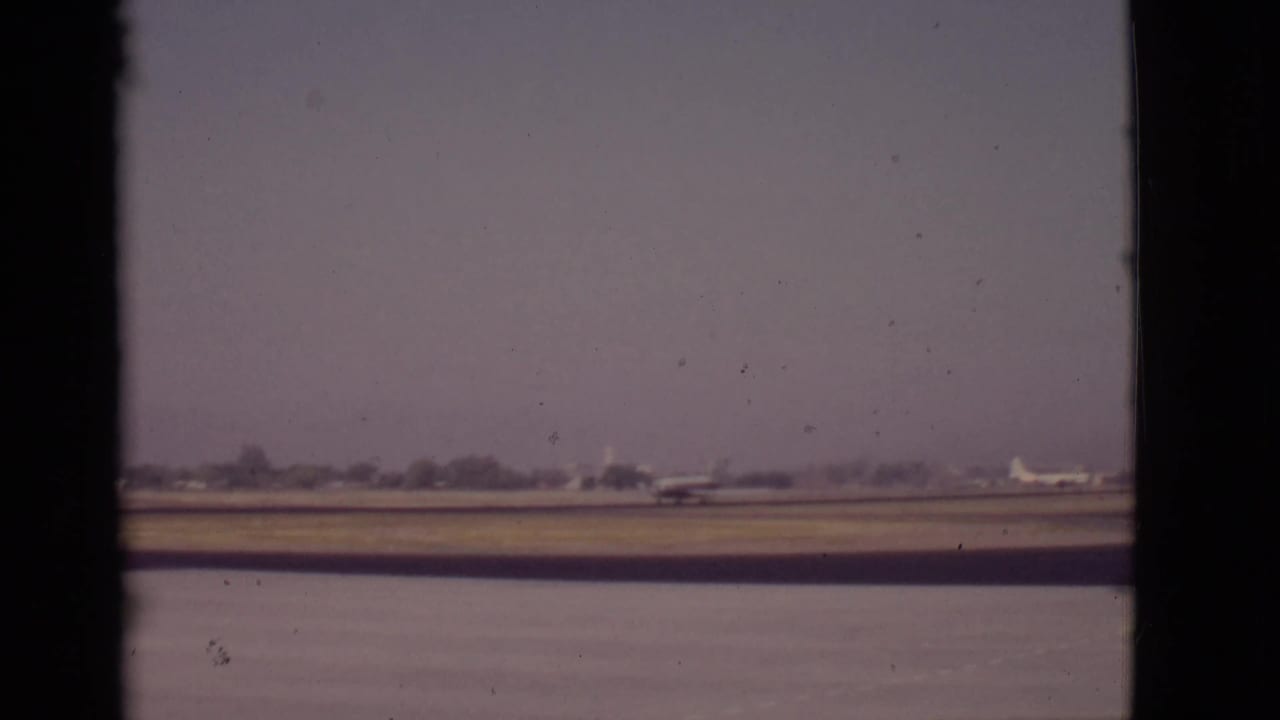 1968 Arizona home-movie frame of a propeller airplane across water on a flat runway, with a hazy treeline in the distance.