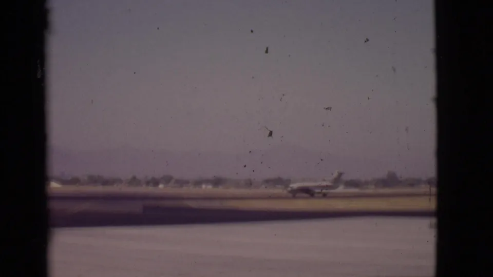 1968 Arizona 8mm frame showing a small passenger aircraft moving along a runway with hazy mountains in the distance.