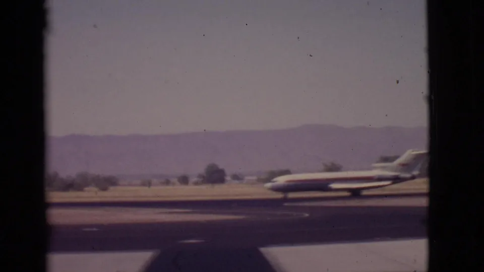 1968 Arizona home-movie frame showing a small passenger aircraft near the right edge of the runway view with mountains behind.