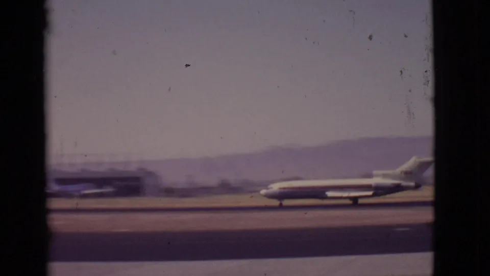 1968 Arizona airport view of a passenger jet taxiing across the runway, seen through dark window framing with haze and distant hills.