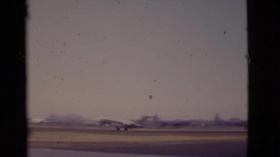 A distant passenger jet travels along the runway in soft 8mm focus, with hazy Arizona mountains and airport structures behind it.