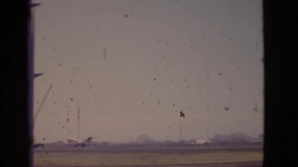 1968 Arizona 8mm frame showing a distant airplane on or near a runway, viewed through dark window mullions in hazy light.