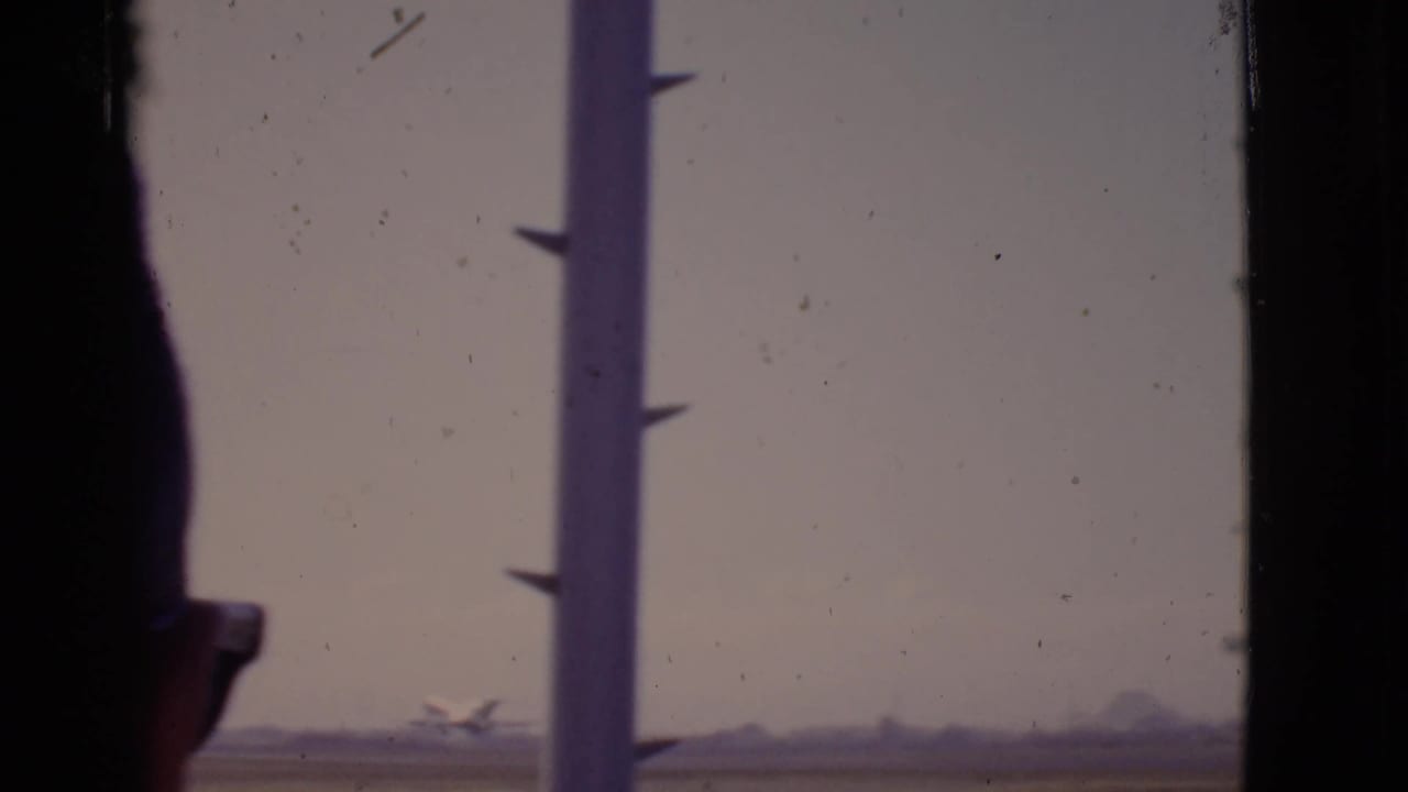 1968 Arizona 8mm view of a small distant airplane taking off, with a vertical airport-window post interrupting the frame.