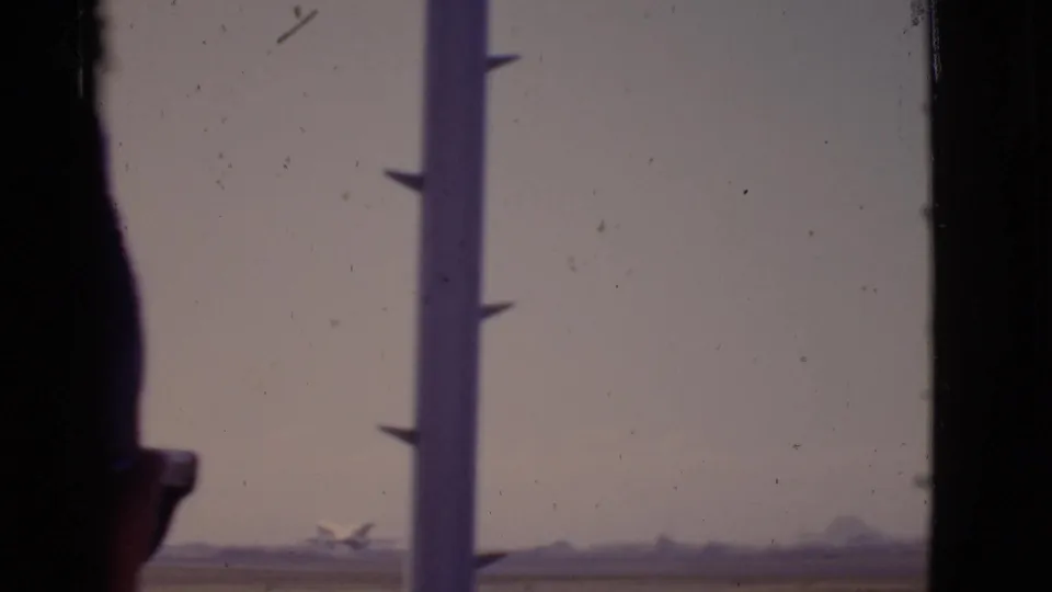 1968 Arizona 8mm view of a small distant airplane taking off, with a vertical airport-window post interrupting the frame.