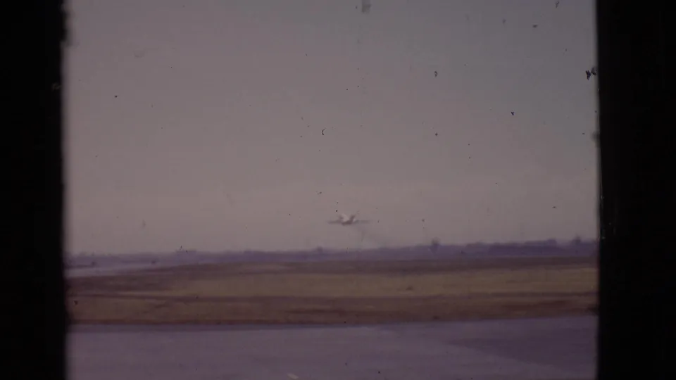 1968 Arizona home-movie frame showing a distant airplane ascending above a flat airport field and runway area.