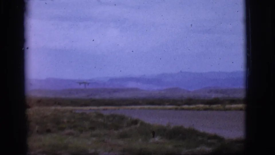 1968 Flagstaff-area 8mm frame with a small jet near the horizon over open ground, water, and blue-toned mountain ridges.