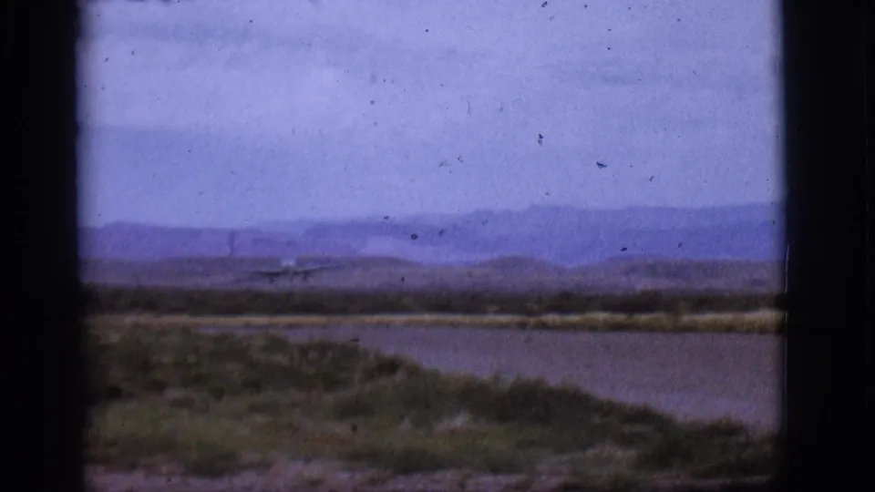 Distant jet nears landing in a wide 1968 8mm view of the Flagstaff, Arizona landscape, with water, scrub, and low mountains behind it.