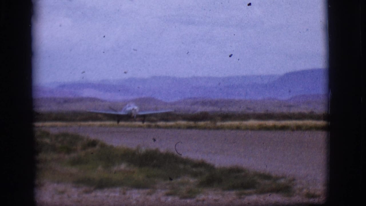 Small blue plane continues along the runway in Flagstaff, Arizona, framed by 8mm gate borders and distant hills.