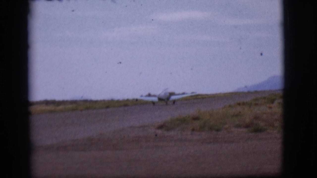 1968 Flagstaff-area home-movie frame showing a small single-engine plane near a runway, seen through dark frame edges with mountains beyond.
