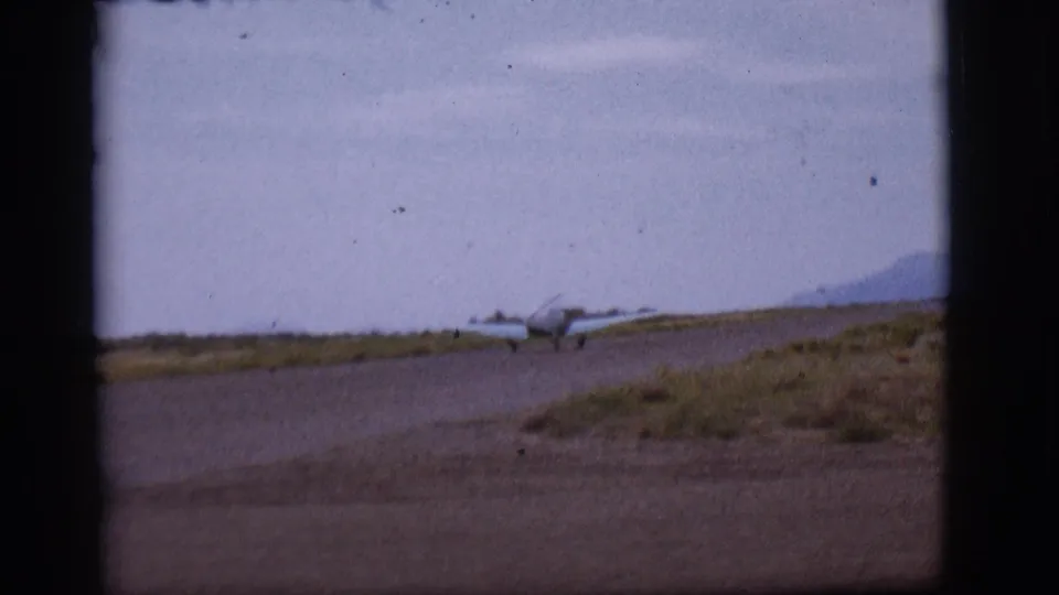 1968 Flagstaff-area home-movie frame showing a small single-engine plane near a runway, seen through dark frame edges with mountains beyond.